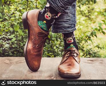 Men's legs in stylish shoes, bright, variegated socks with Christmas and New Year's patterns on the wooden terrace on the background of green trees. Beauty, fashion, elegance. Men's legs in stylish shoes, bright, variegated socks