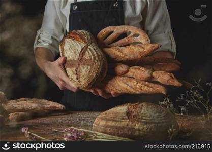 Men’s hands hold a variety of bread on the black background of a wooden table with dry flowers. Variety of bread hold men’s hands