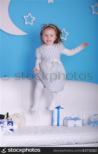 Merry Christmas and happy holidays. New Year 2021. portrait of a little girl on a white sofa with Christmas presents. New Year holidays concept. blue wall with a white moon on a background.. Merry Christmas and happy holidays. New Year 2021. portrait of a little girl on a white sofa with Christmas presents. New Year holidays concept. blue wall with a white moon on a background