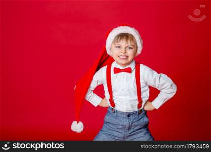 Merry Christmas. Cute cheerful cunning little boy in Santa Claus hat on red background. A happy childhood with dreams and gifts. Close-up of baby&rsquo;s open mouth, milk tooth fell out.. Merry Christmas. Cute cheerful little boy in Santa Claus hat on red background.