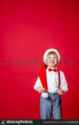 Merry Christmas. Cute cheerful little boy in Santa Claus hat on red background. A happy childhood with dreams and gifts. Close-up of baby&rsquo;s open mouth, milk tooth fell out.. Merry Christmas. Cute cheerful little boy in Santa Claus hat on red background.