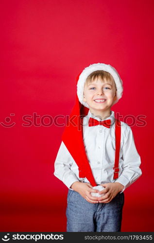 Merry Christmas. Cute cheerful little boy in Santa Claus hat on red background. A happy childhood with dreams and gifts. Close-up of baby&rsquo;s open mouth, milk tooth fell out.. Merry Christmas. Cute cheerful little boy in Santa Claus hat on red background.