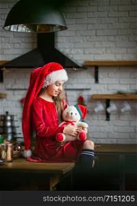 Merry Christmas. Cute little girl in Santa hat is holding a teddy bear sitting in the kitchen, waiting for the holiday. A time of miracles and fulfillment of desires.. Merry Christmas. Cute little girl in Santa hat is holding a teddy bear sitting in the kitchen, waiting for the holiday.