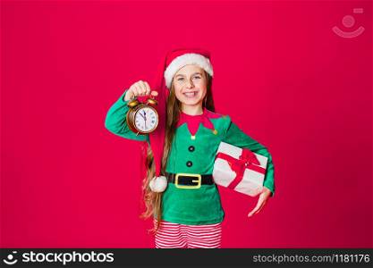 Merry Christmas, happy attractive girl with a clock and gifts in the costume of Santa Claus helper on a bright red bright color background. Portrait of a beautiful elven baby. Copy space.. Merry Christmas, happy attractive girl with a clock and gifts in the costume of Santa Claus helper on a bright red bright color background.