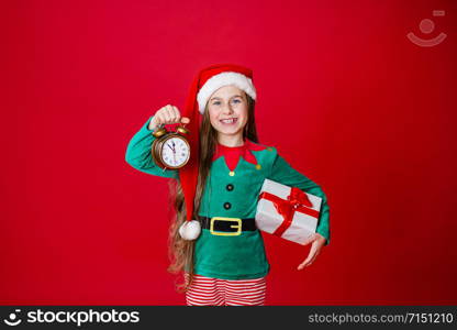 Merry Christmas, happy attractive girl with a clock and gifts in the costume of Santa Claus helper on a bright red bright color background. Portrait of a beautiful elven baby. Copy space.. Merry Christmas, happy attractive girl with a clock and gifts in the costume of Santa Claus helper on a bright red bright color background.