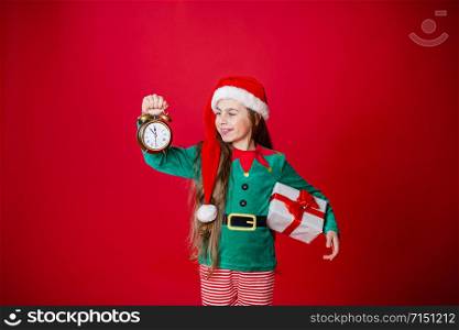 Merry Christmas, happy attractive girl with a clock and gifts in the costume of Santa Claus helper on a bright red bright color background. Portrait of a beautiful elven baby. Copy space.. Merry Christmas, happy attractive girl with a clock and gifts in the costume of Santa Claus helper on a bright red bright color background.