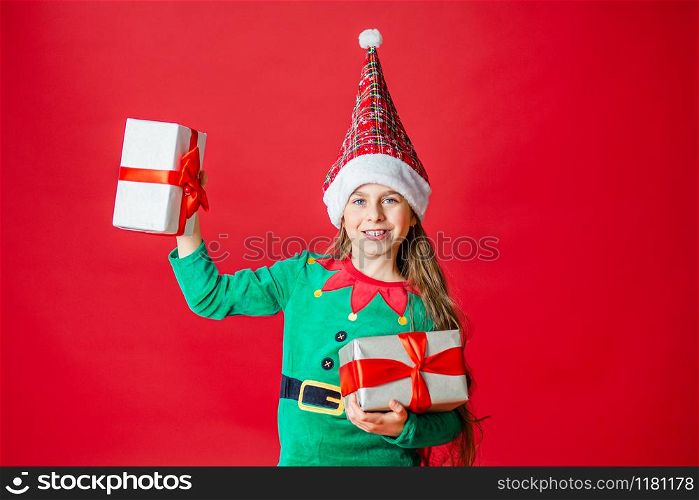 Merry Christmas, happy attractive girl with gifts in a costume of Santa Claus helper elf on a bright red bright color background. Portrait of a beautiful elven baby. Copy space.. Merry Christmas, happy attractive girl with gifts in a costume of Santa Claus helper elf on a bright red bright color background.