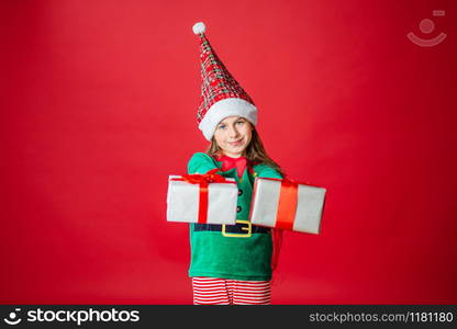 Merry Christmas, happy attractive girl with gifts in a costume of Santa Claus helper elf on a bright red bright color background. Portrait of a beautiful elven baby. Copy space.. Merry Christmas, happy attractive girl with gifts in a costume of Santa Claus helper elf on a bright red bright color background.