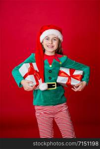 Merry Christmas, happy attractive girl with gifts in a costume of Santa Claus helper elf on a bright red bright color background. Portrait of a beautiful elven baby. Copy space.. Merry Christmas, happy attractive girl with gifts in a costume of Santa Claus helper elf on a bright red bright color background.