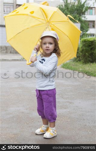 Merry little girl stands with the yellow umbrella on an asphalt road on the street