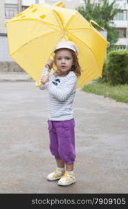 Merry little girl stands with the yellow umbrella on an asphalt road on the street