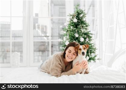 Merry lovely woman with cute dog, embraces pet and expresses love, dressed in winter sweater, pose together at comfortable bed in white spacious room, decorated firtree behind for festive mood