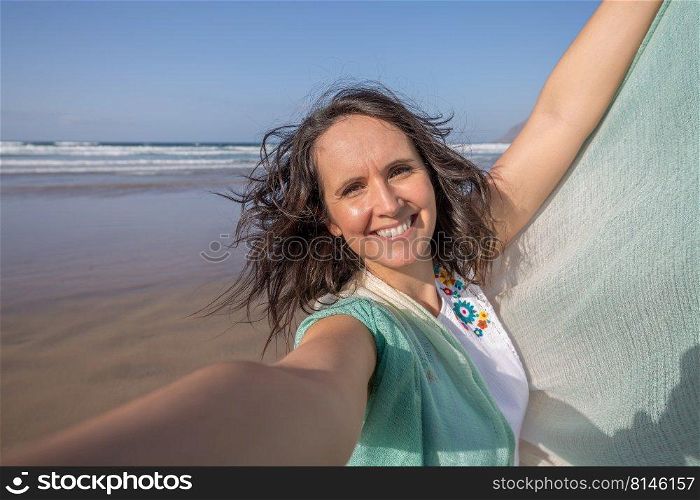 Merry middle aged female tourist with dark hair raising arm with scarf and looking at camera with smile while taking selfie against waving sea and blue sky during summer vacation. Woman taking selfie near sea