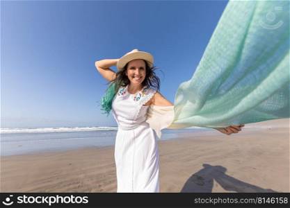 Merry middle aged woman in white dress with scarf flying on wind touching straw hat and looking at camera with smile while spending time on sandy beach near sea on cloudless summer day. Happy female traveler resting on beach