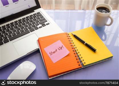 Message on paper with text Start Up ! stick on colorful book with laptop and a cup of coffee on glass table, top view image