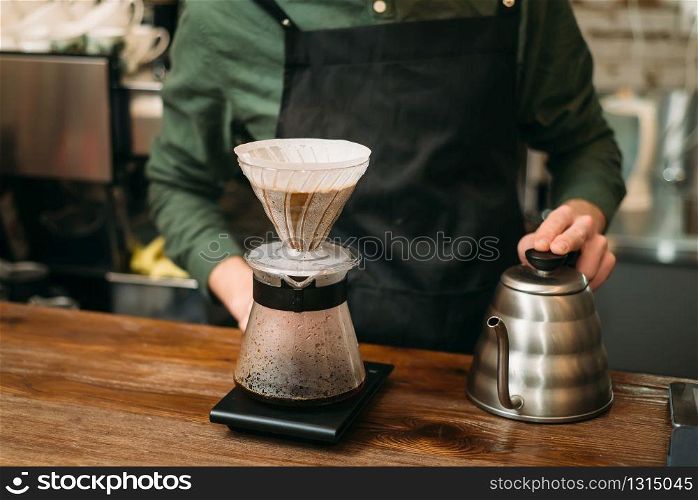 Metal coffee pot and glass on a bar counter against waiter in black apron. Blury coffee house on background.. Metal coffee pot and glass on a bar counter.