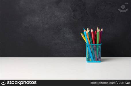metal glass with pens, pencils and felt-tip pens and scissors on the background of an empty black chalk board, white table, copy space