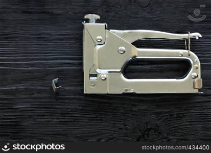 Metal industrial chrome stapler and brackets to it lie on a black wooden table with the texture of old wood.. Metal industrial chrome stapler gun and staples to it on a black background