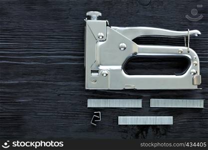 Metal industrial chrome stapler and cassette brackets to it lie on a black wooden table with the texture of old wood.. Metal industrial chrome stapler gun and staples to it on a black background