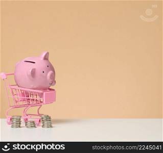 metal miniature shopping cart and pink piggy bank on a white table, brown background. Concept of saving budget, discounts, copy space