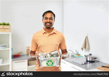 metal recycling, waste sorting and sustainability concept - smiling young indian man in striped t-shirt holding plastic box with tin cans over home kitchen background. smiling young indian man sorting metallic waste