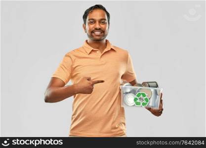 metal recycling, waste sorting and sustainability concept - smiling young indian man in striped t-shirt holding plastic box with tin cans over grey background. smiling young indian man sorting metallic waste