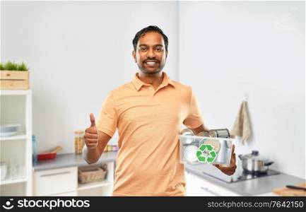 metal recycling, waste sorting and sustainability concept - smiling young indian man holding plastic box with tin cans showing thumbs up over home kitchen background. smiling young indian man sorting metallic waste