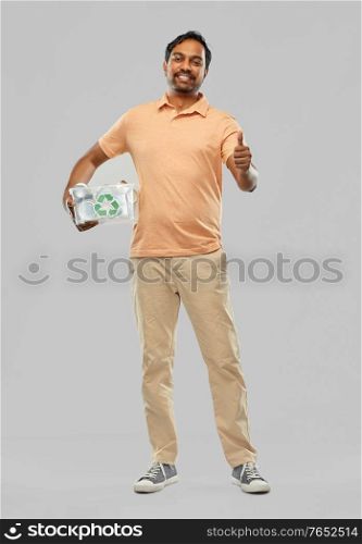 metal recycling, waste sorting and sustainability concept - smiling young indian man in striped t-shirt holding plastic box with tin cans and showing thumbs up over grey background. smiling young indian man sorting metallic waste