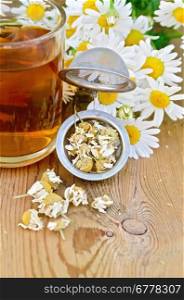 Metal strainer with dried chamomile, a bouquet of fresh flowers chamomile, tea in glass mug on a background of wooden boards