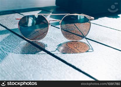 Metal thin trendy sunglasses of brown tint, on wooden table. Sunny day, outdoors, cute reflection and shade. Closeup image, minimal style. Copy space for your text and design. Lounge, travel concept