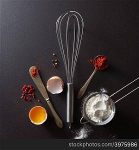 Metal whisk, spoon with red pepper knife and halved raw egg on a black concrete background with copy space. Cooking Concept. Flat lay. Spoon with red pepper, metal whisk, vintage knife raw egg and flour on a black concrete background with copy space. Concept cooking. Flat lay