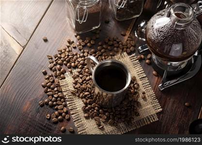 Metallic cup and vacuum coffee maker also known as vac pot, siphon or syphon coffee maker and toasted coffee beans on rustic wooden table