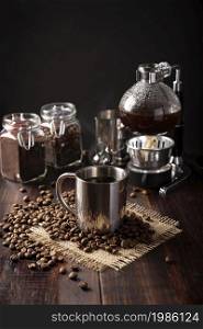 Metallic cup and vacuum coffee maker also known as vac pot, siphon or syphon coffee maker and toasted coffee beans on rustic wooden table