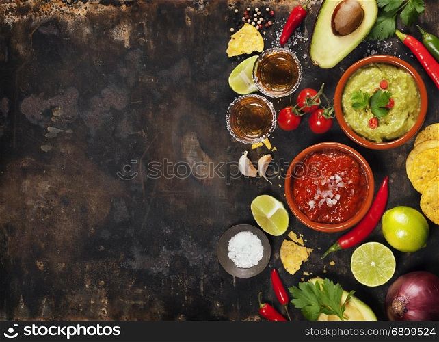 Mexican food concept: tortilla chips, guacamole, salsa, tequila shots and fresh ingredients over vintage rusty metal background. Top view