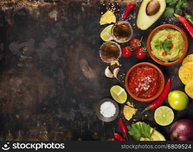 Mexican food concept: tortilla chips, guacamole, salsa, tequila shots and fresh ingredients over vintage rusty metal background. Top view