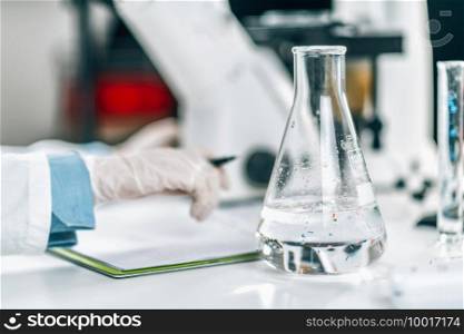 Microplastic Pollution Analysis. Environment pollution research scientist taking notes in a laboratory, flask with water s&le in focus. Microplastic Pollution Analysis. 