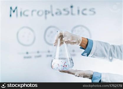 Microplastic pollution concept. Scientist holding a flask with a water sample full of tiny plastic particles against a whiteboard with microplastic pollution graph.. Microplastic Pollution Concept