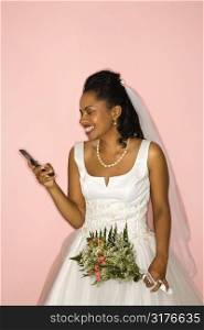 Mid-adult African-American bride looking at cellphone on pink background.