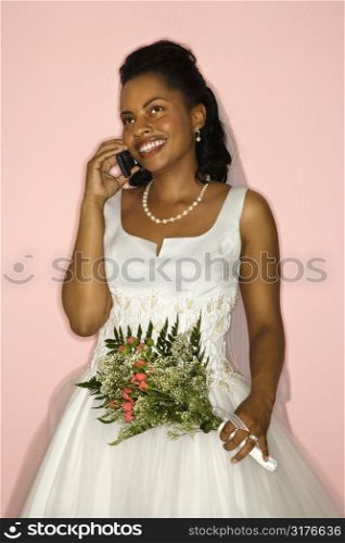 Mid-adult African-American bride talking on cellphone on pink background.