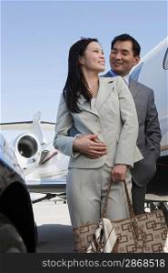 Mid-adult Asian business couple standing in front of car and airplane.