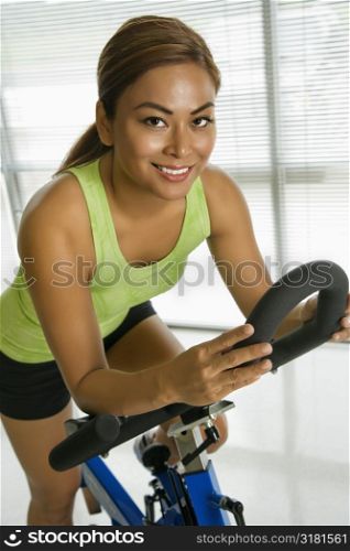 Mid adult Asian woman pedaling exercise bicycle smiling at viewer.