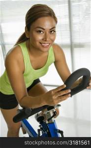Mid adult Asian woman pedaling exercise bicycle smiling at viewer.