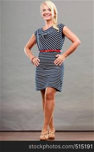 Mid adult blond attractive woman in full length wearing striped dress sandals shoes posing, studio shot on gray. Mid adult woman in summer striped dress
