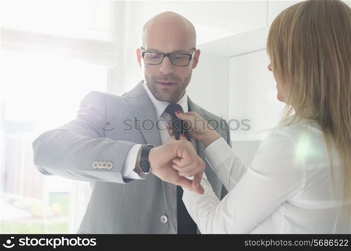 Mid adult businessman checking wristwatch while woman adjusting his tie at home