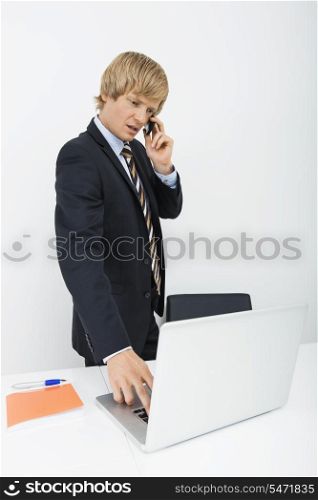 Mid adult businessman conversing on cell phone while using laptop in office