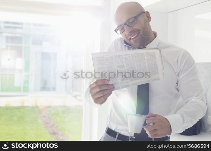 Mid adult businessman on call while holding newspaper and coffee cup at home