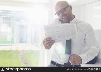 Mid adult businessman on call while holding newspaper and coffee cup at home