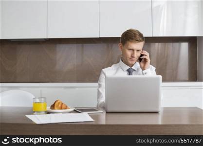 Mid adult businessman on call while using laptop at breakfast table