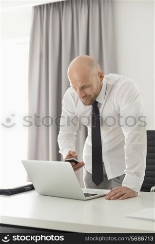 Mid adult businessman using call phone and laptop at table in home office