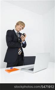Mid adult businessman using cell phone at desk with laptop in office